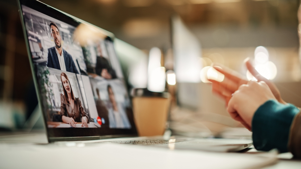 A person gestures during a video conference call with four colleagues shown on a laptop screen