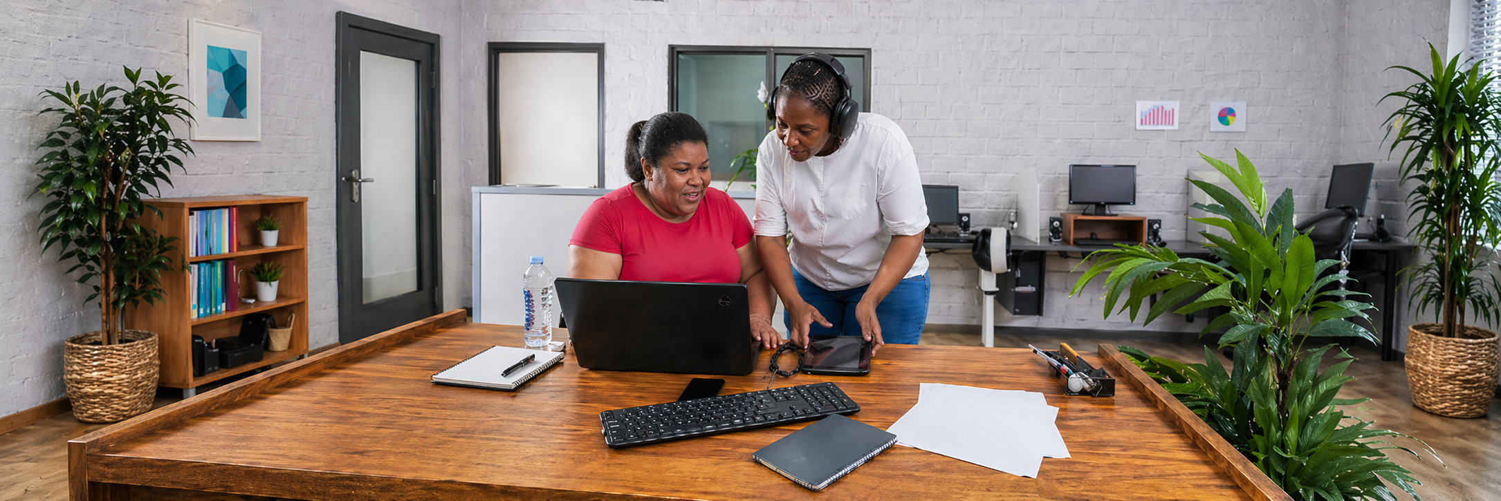 A visually impaired woman and a colleague work together at a desk in a bright office, using a laptop and tablet.